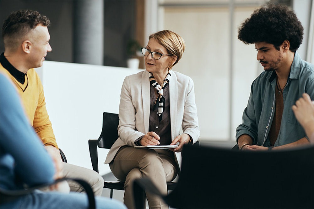 Middle-aged therapist with glasses discusses addiction and mental health dual diagnosis treatment with two young men in group therapy