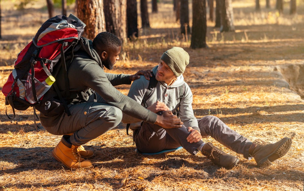 Man wearing Timberland boots and carrying hiking backpack reassuring sitting man with hiking backpatch clutching shin