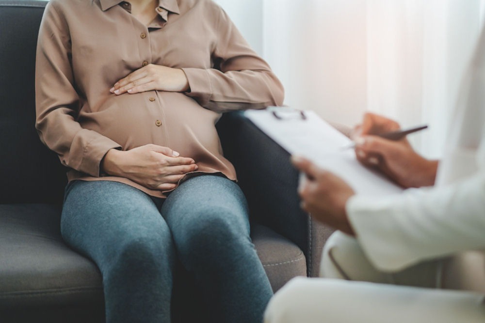 Woman embracing pregnant belly under brown button-up shirt while talking with seated therapist holding clipboard