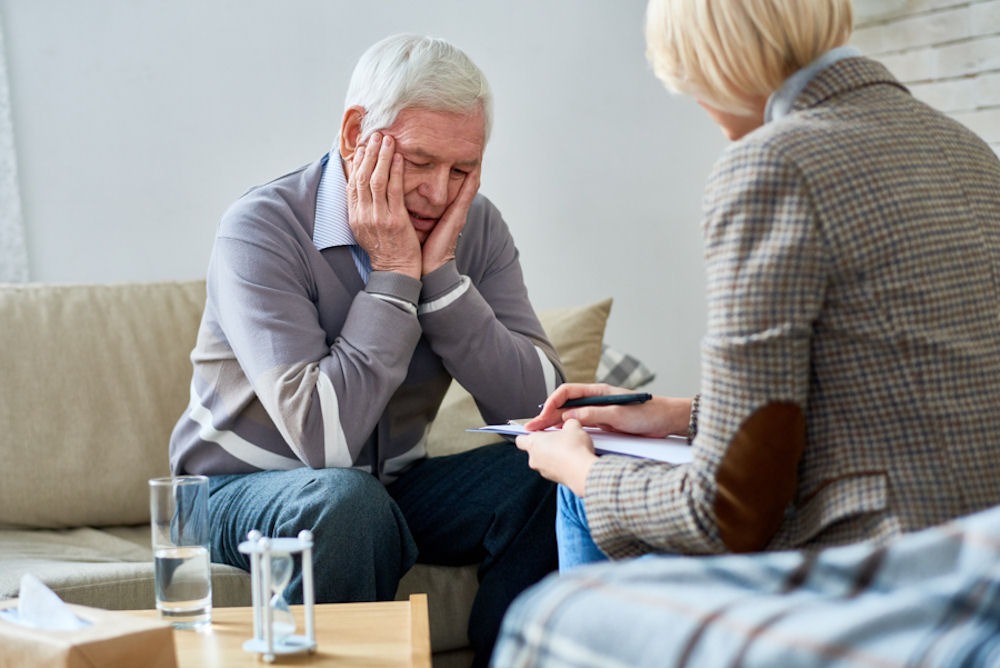 Older man holds head in hands while talking about sobriety to therapist taking notes