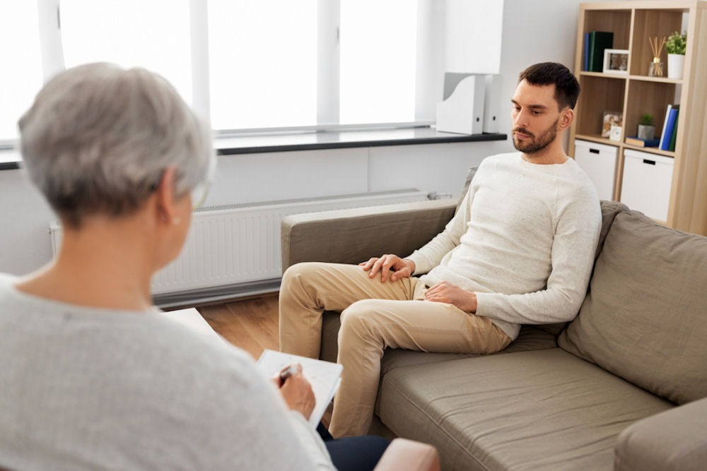 Uneasy man on couch talking to addiction therapist as she takes notes