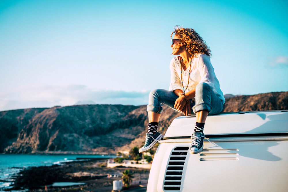 Woman sitting on top of a small white bus, looking out onto the ocean from the beach