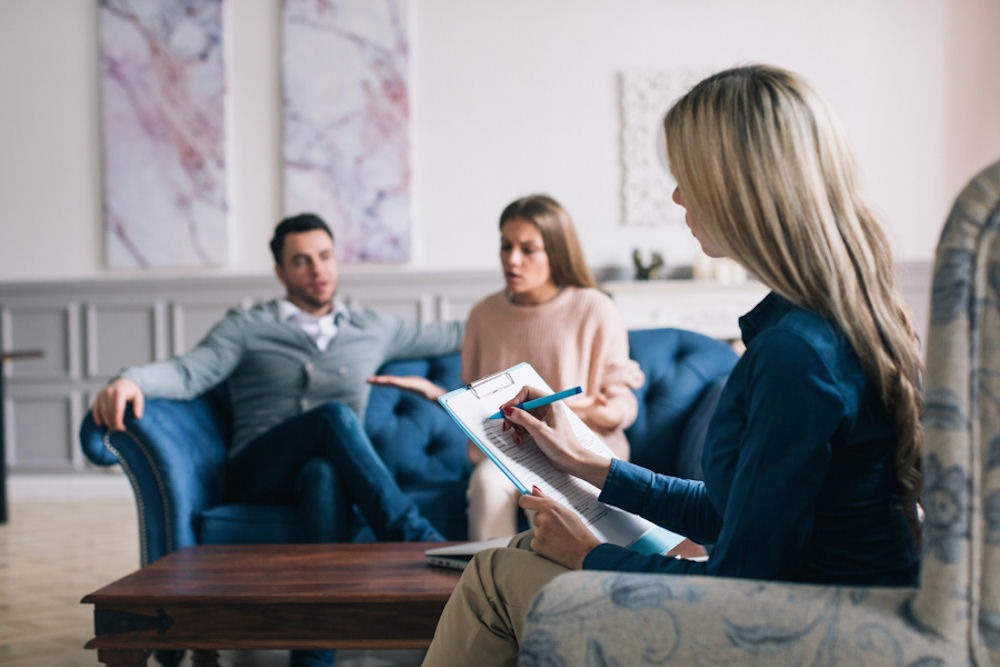 Young couple arguing on blue couch while therapist takes notes