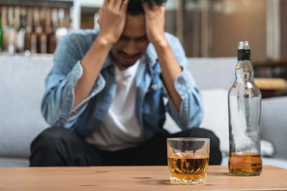 Frustrated man sitting on grey couch in front of tall glass of whiskey neat