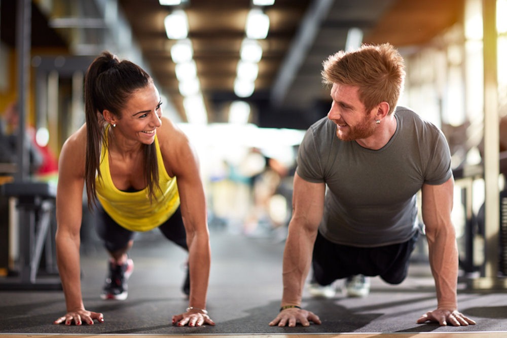 Man and woman in gym smiling at each other while performing plank pose