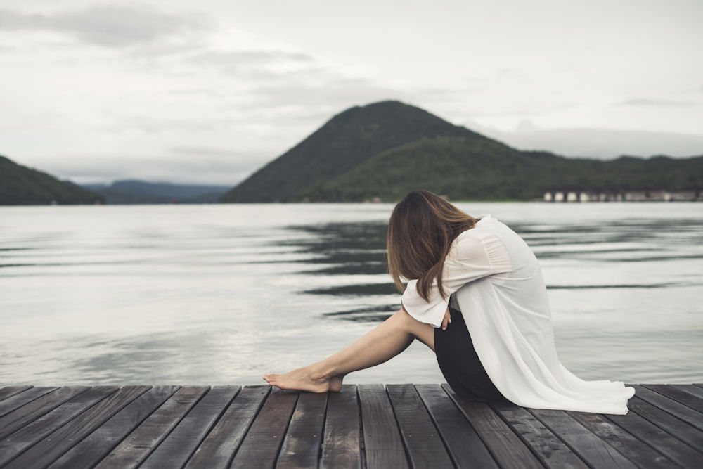 Woman slumped over and hiding face while sitting on a wooden dock on a lake