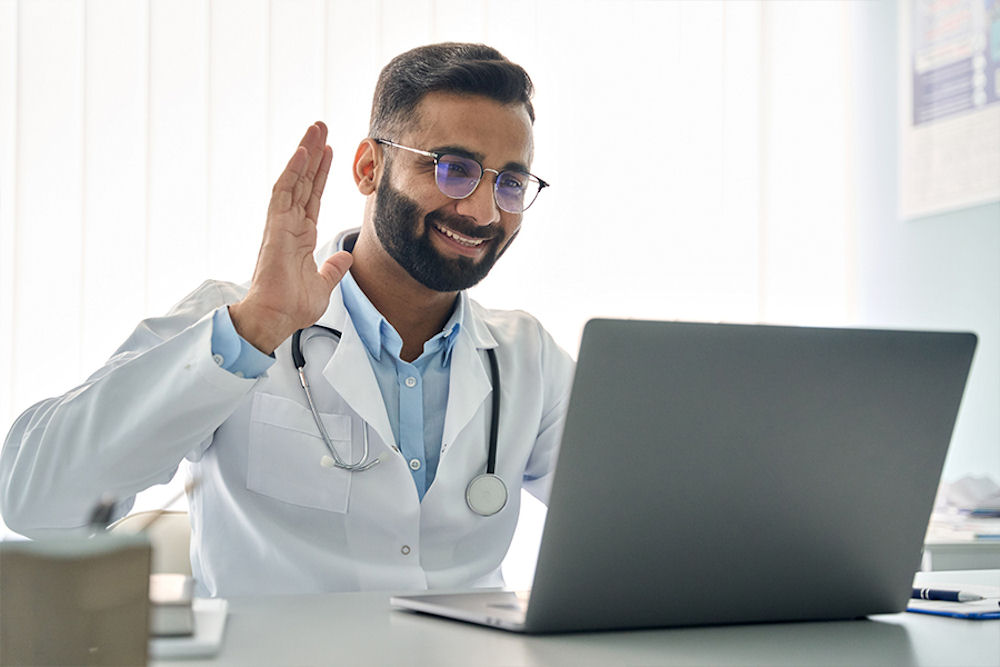 Doctor with glasses and stethoscope saying hello to someone he's communicating with over telehealth rehab program