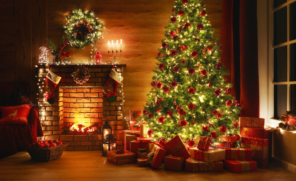 Wide shot of Christmas tree with wrapped presents underneath, next to lit fireplace with decorated mantle below lit wreath