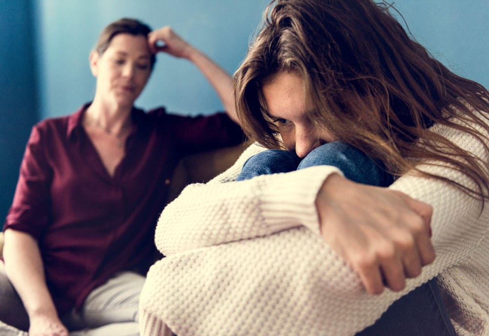 Woman in a waffle knit shirt hugging knees while disapproving mother looks on