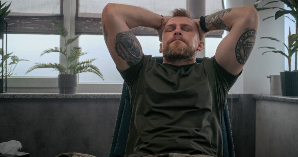 Man with tattoos relaxing in inner space with plant after discussing a higher power in recovery treatment