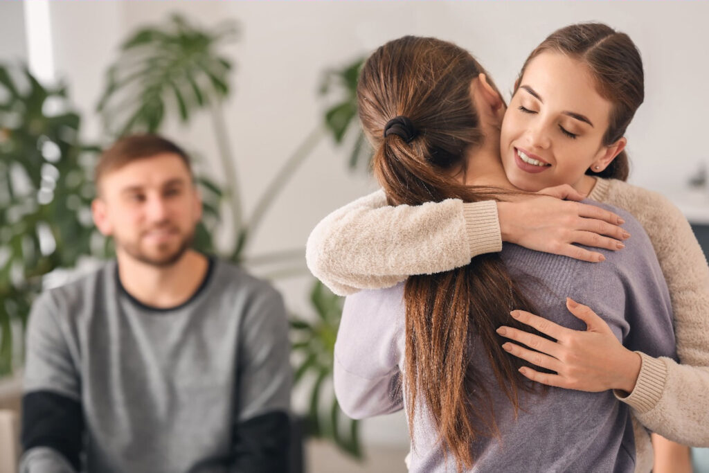 Young woman embracing another woman in depression support group as smiling man looks on