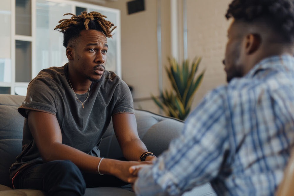 Heristant young man with dreadlocks learning about urge surfing from another man on a grey couch.