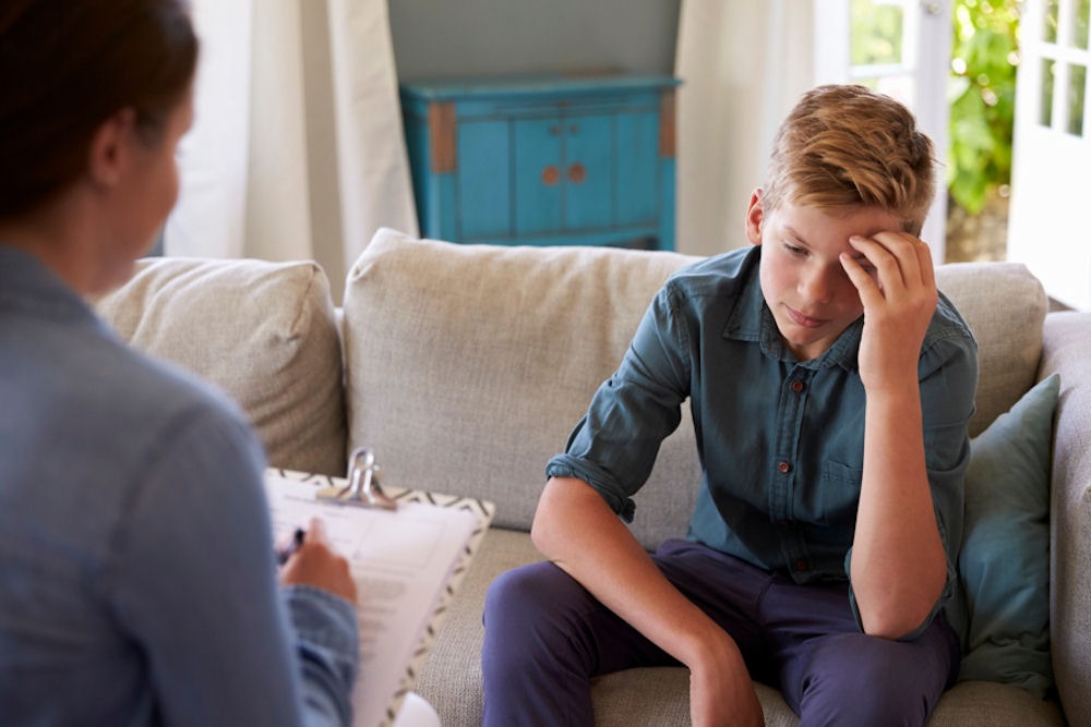 Teenage boy holding head as therapist takes notes.