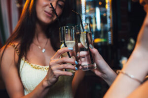 Two women clinking glasses of water with lemon served in highball glasses.