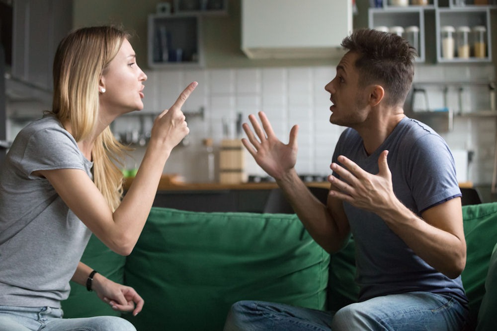Man and woman arguing on a couch with kitchen in the background.