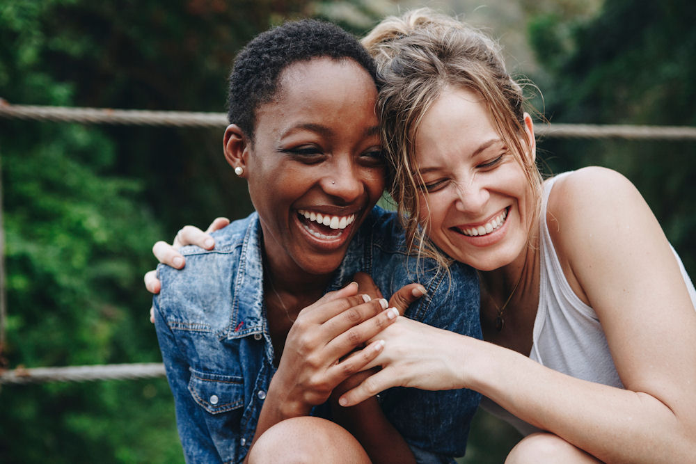 Girlfriends embrace playfully while sitting on rope bridge.