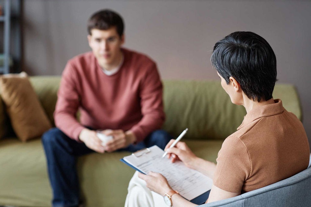Young man talking about an anxiety disorder with a therapist holding a clipboard.